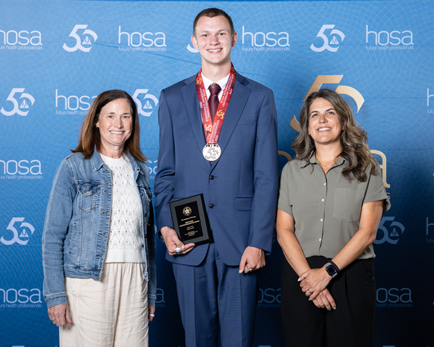 Three people stand in front of a HOSA banner. One of them wears a medal and holds an award plaque.