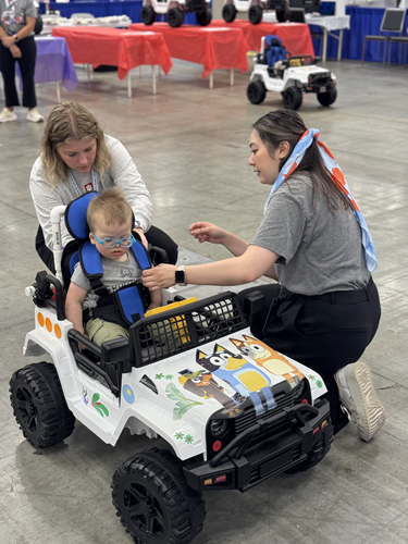 A young child sits in an adaptive car decorated with Bluey characters while two adults strap the child in.
