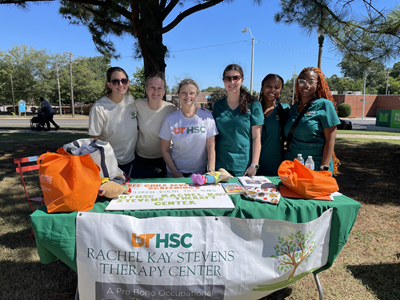 Six women stand outside behind a table. Attached to the table's front is a banner that says UT HSC Rachel Kay Stevens Therapy Center.