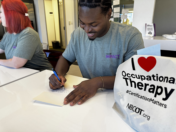 A smiling man writes a note in a card. A tote bag is next to him printed with "I heart symbol Occupational Therapy; #CertificationMatters; NBCOT.org"