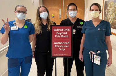 Three women and one man wearing masks and scrubs stand in a hallway behind a sign: COVID unit beyond this point. Authorized personnel only.