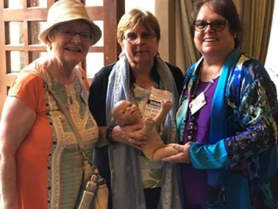 Three women smile at the camera. Two of the women hold a baby doll.