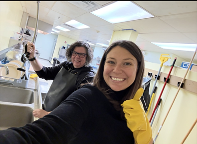 Two women in a kitchen. One woman makes a thumbs up gesture, the other holds a faucet head.