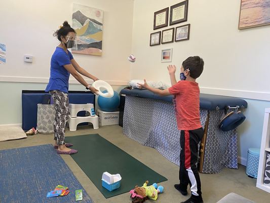 Dr. Smith holds a toilet seat while a young boy tosses a ball-shaped object that is aimed toward the toilet seat.